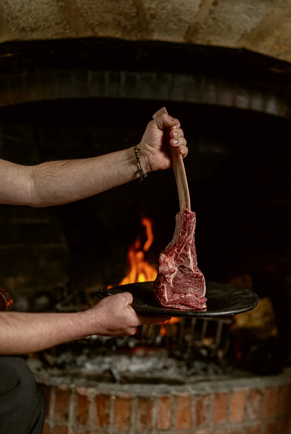 Mesero presentando un corte tomahawk junto al fuego del horno de leña